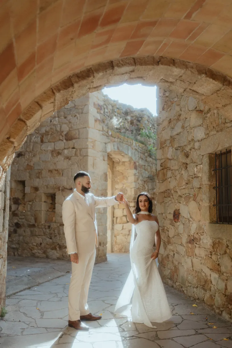 A couple dances beneath a sunlit stone archway, celebrating their surprise proposal in a historic village setting.