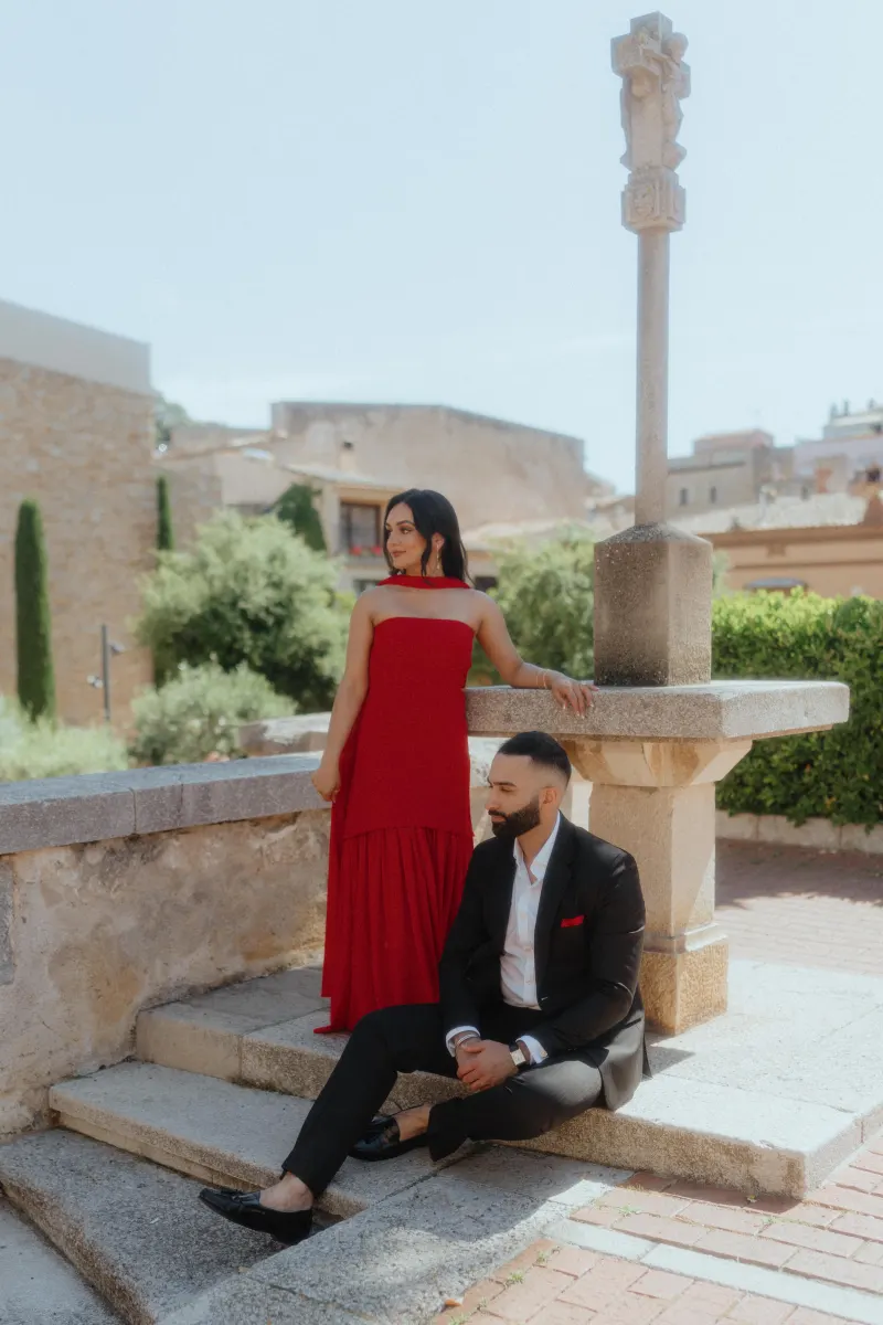 A couple dressed elegantly in red and black share a quiet moment by a stone monument in Begur after a proposal.