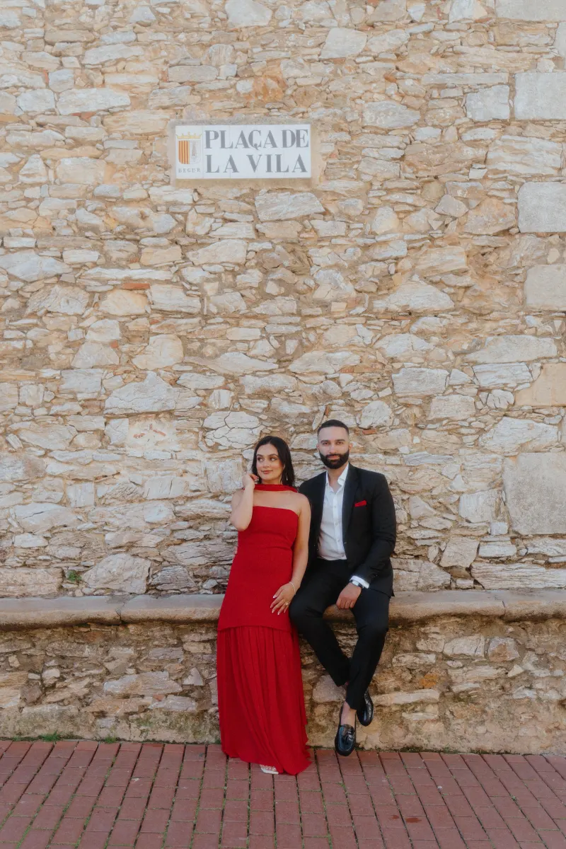 A couple dressed elegantly in red and black share a quiet moment on a stone bench during a surprise proposal shoot.