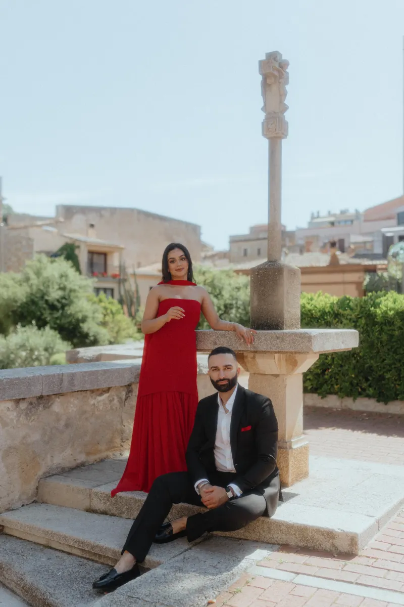 A couple dressed elegantly poses by a historic stone cross in Begur, marking their surprise proposal photoshoot.