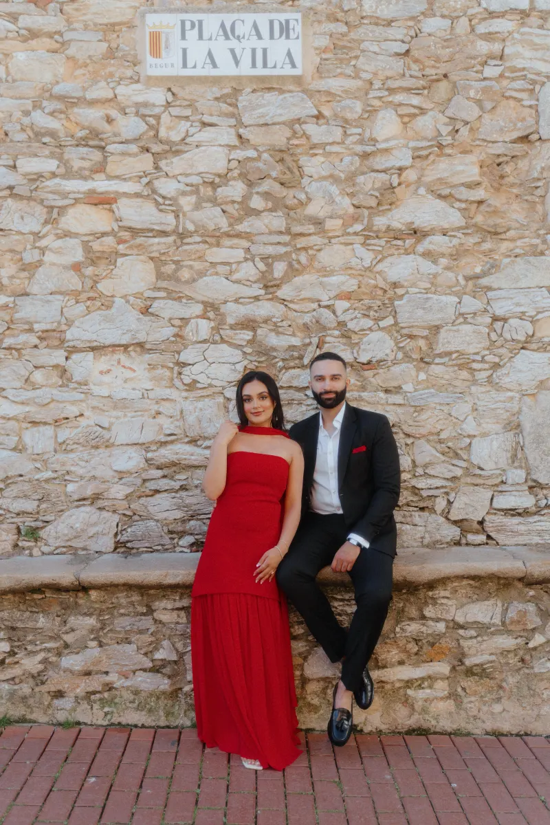A couple dressed elegantly sits by a rustic stone wall in Begur, captured during their surprise proposal photoshoot.
