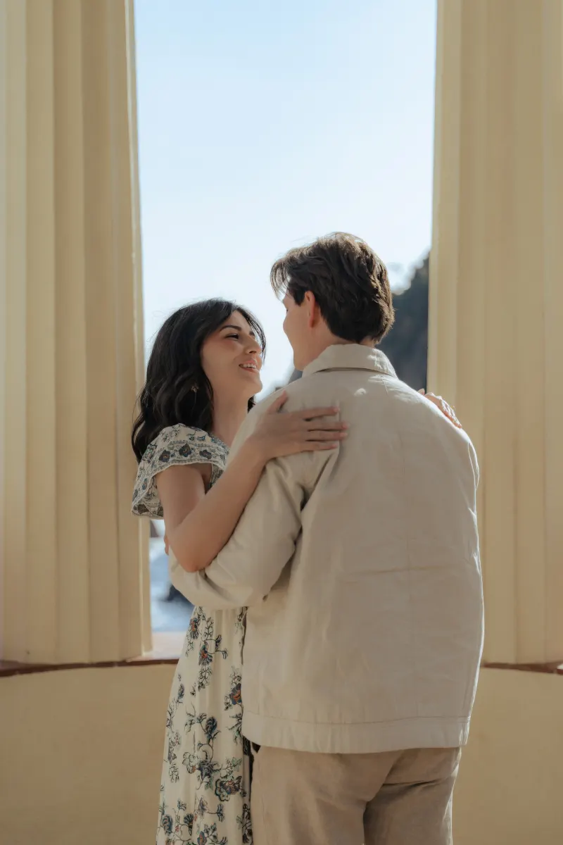 A couple embraces and smiles beneath sunlit columns during a seaside surprise proposal photoshoot.