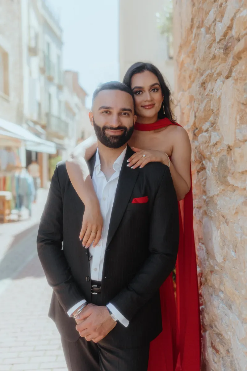 A couple embraces and smiles by a sunlit stone wall in Begur, celebrating their surprise proposal photoshoot.