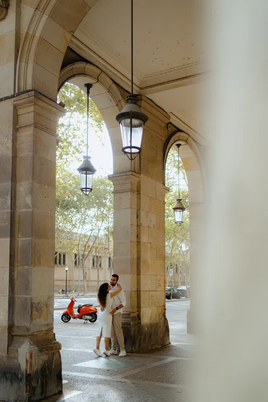 A couple embraces beneath grand stone arches during a surprise proposal photoshoot in a European city street.
