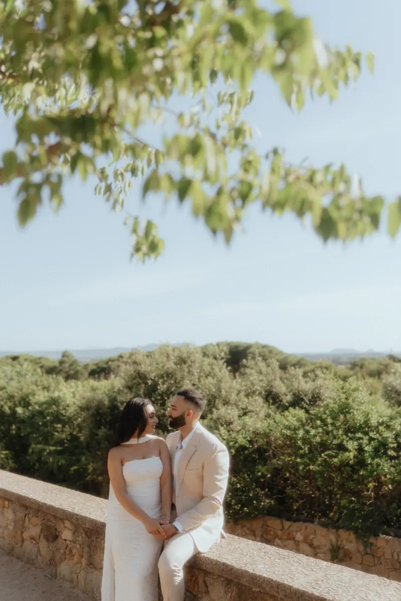 A couple gazes at each other, holding hands on a sunlit stone wall after their surprise proposal photoshoot.