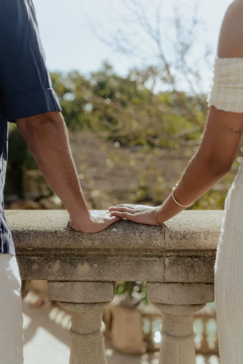 A couple gently touching hands on a sunlit stone railing, capturing a quiet connection during an engagement session.
