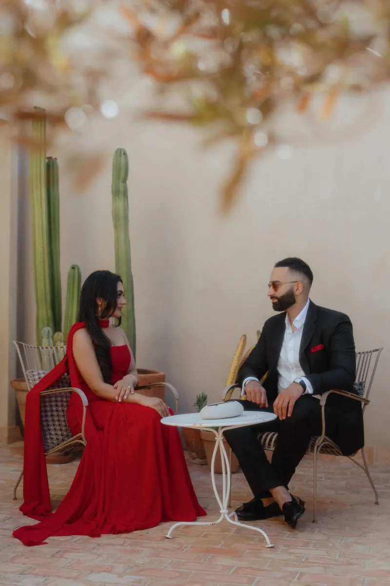 A couple in elegant attire shares a tender moment at a patio table, captured during a surprise proposal photoshoot.