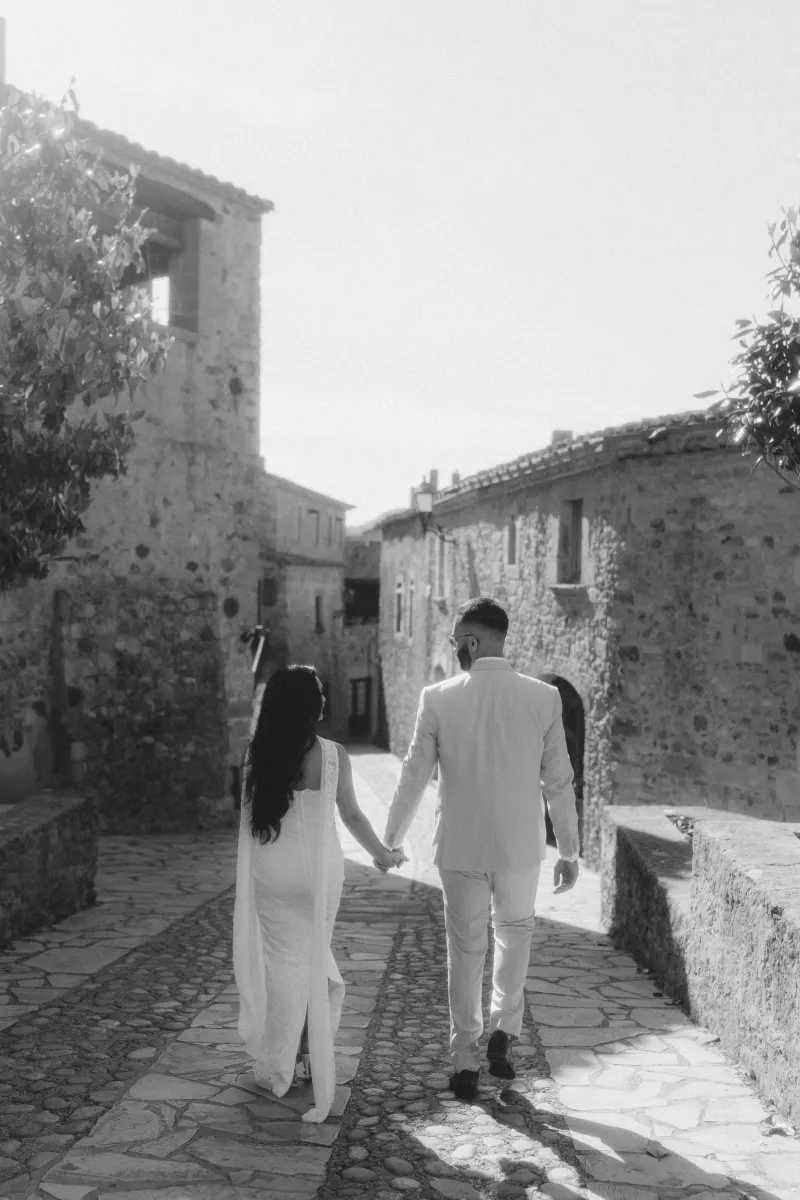 A couple in elegant attire walks hand in hand through a charming stone village after their surprise proposal session.