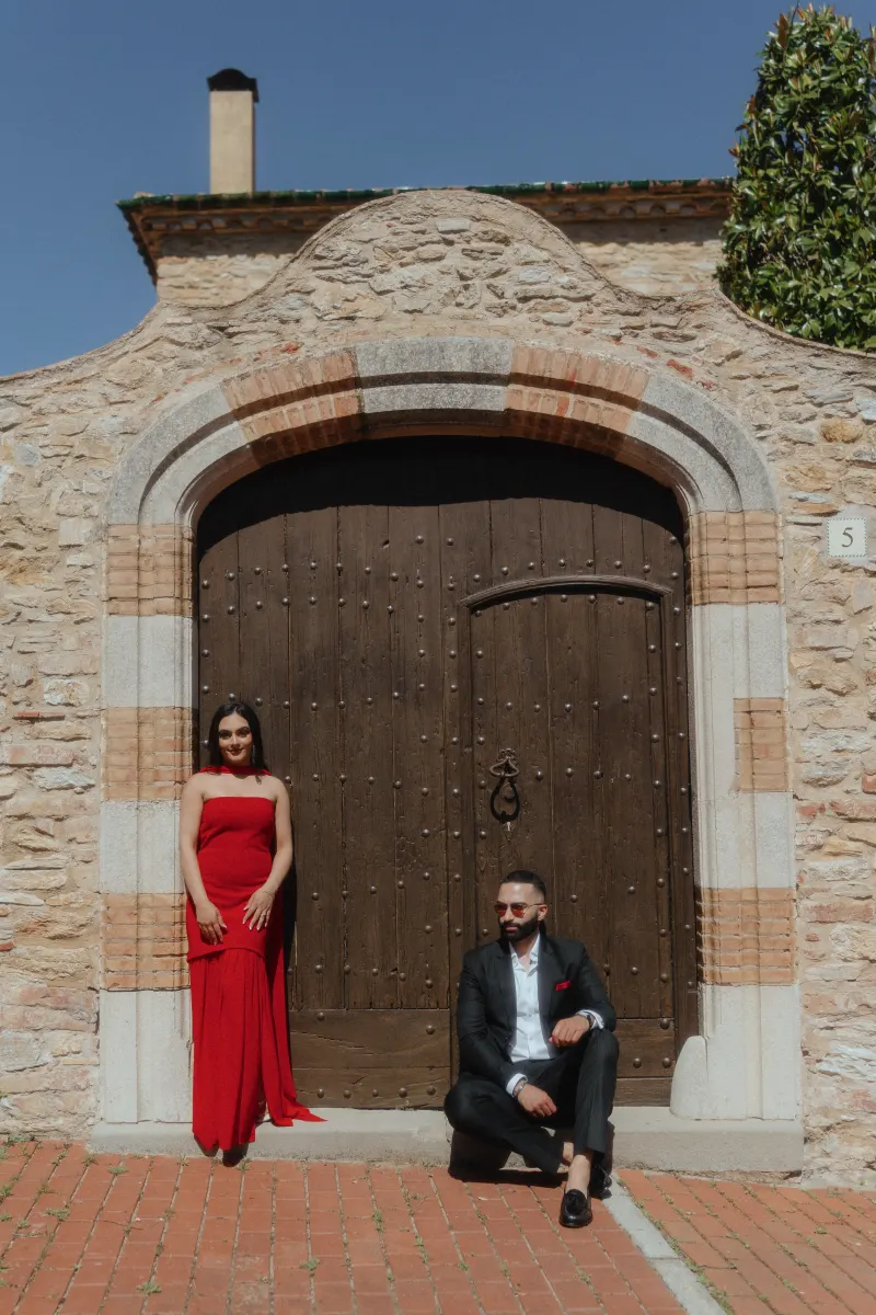 A couple in elegant outfits poses by a rustic wooden door in Begur during their surprise proposal photoshoot.