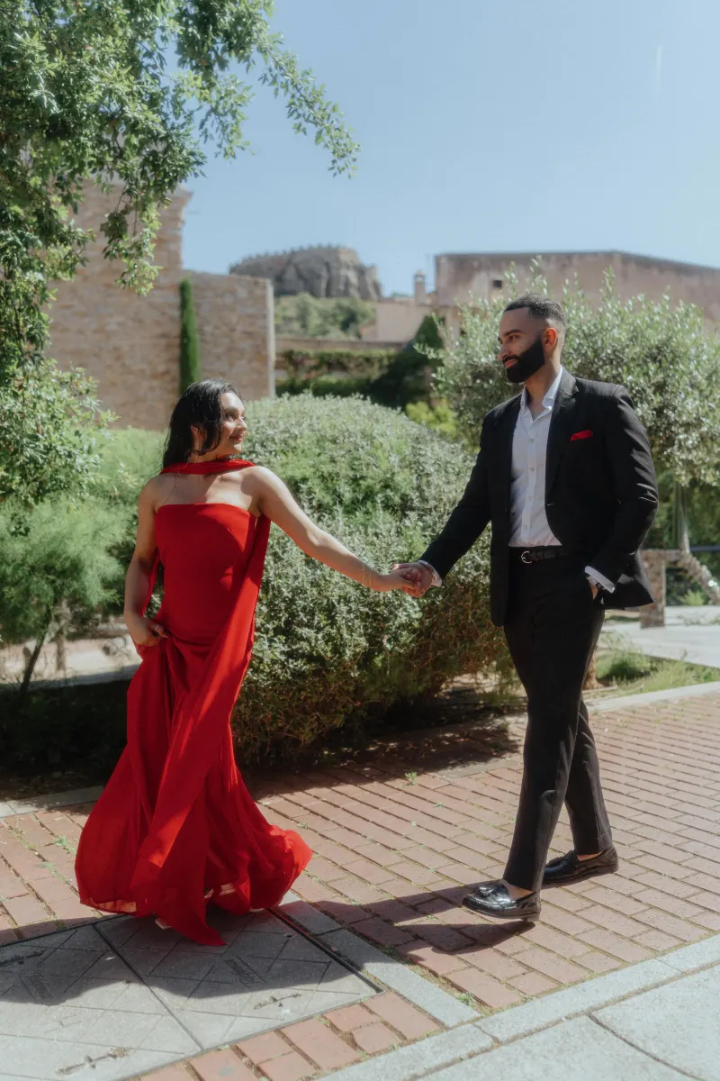 A couple in formal attire shares a joyful moment, holding hands in a sunlit Begur garden during a surprise proposal session.