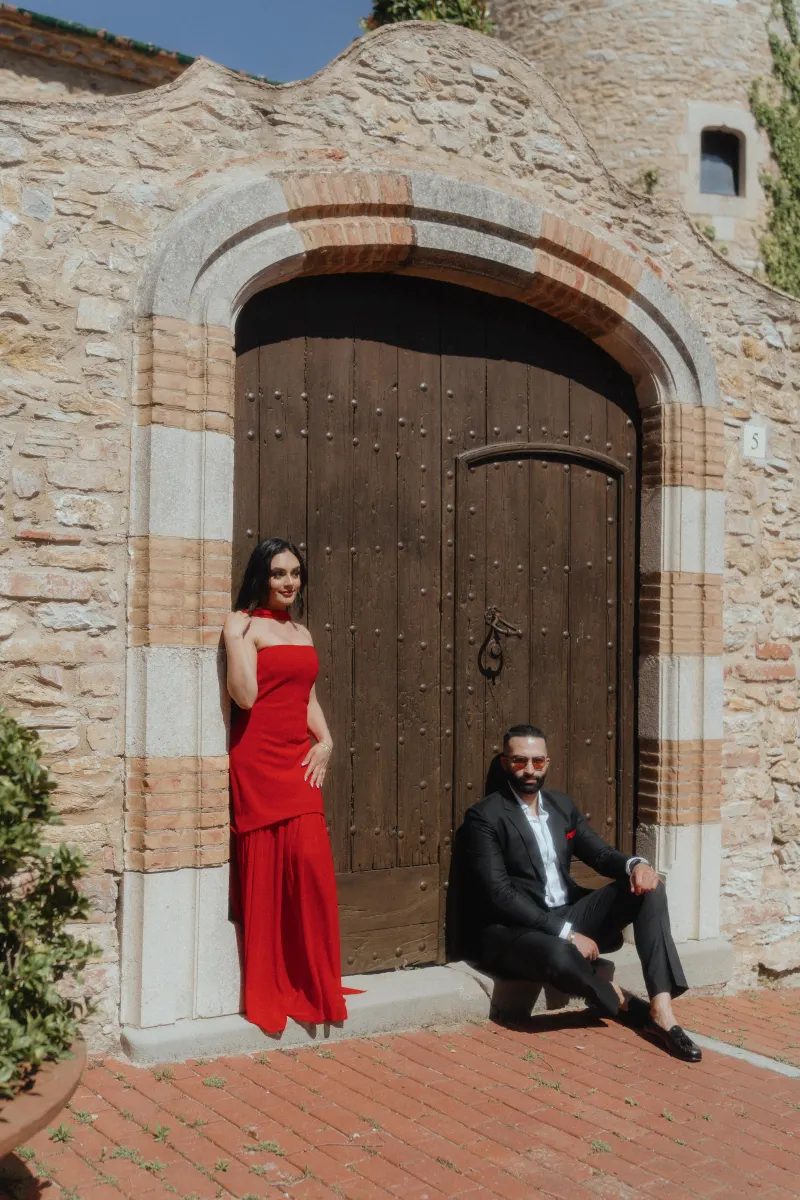 A couple in stylish formalwear poses by a historic wooden door in Begur during their surprise proposal photoshoot.