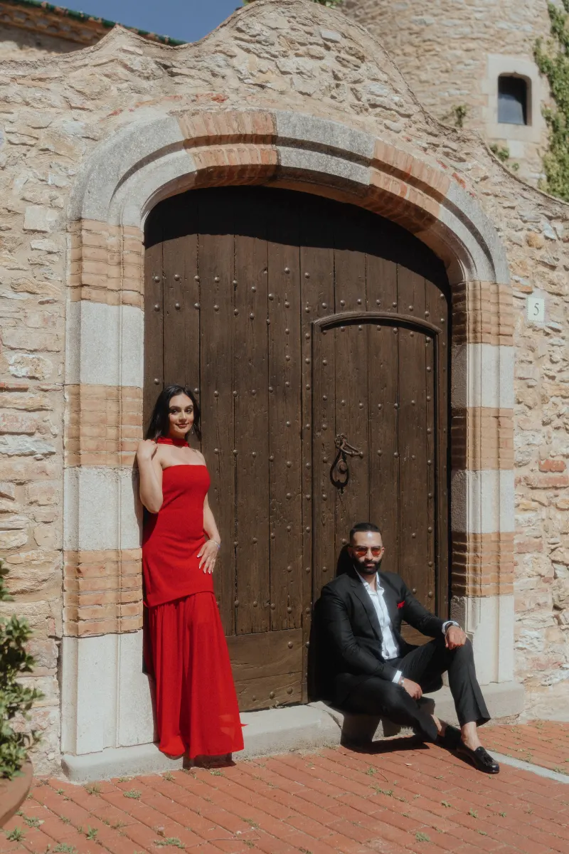 A couple in stylish red and black attire poses by a historic wooden door in Begur during their surprise proposal session.