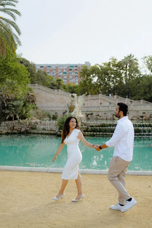 A couple laughs and holds hands by the turquoise fountain during a surprise proposal photoshoot at Parc de la Ciutadella.