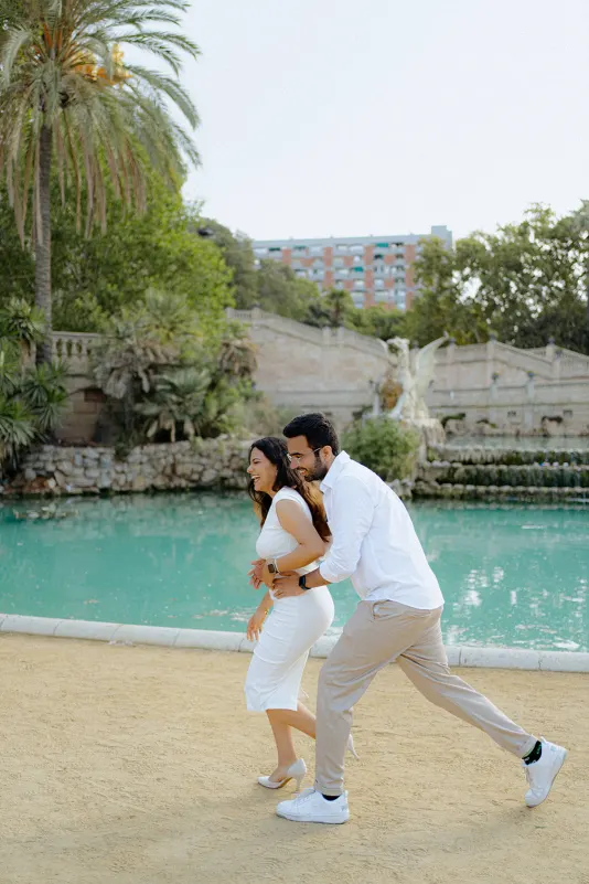 A couple laughs and playfully runs by the turquoise water during a surprise proposal photoshoot at Parc de la Ciutadella.