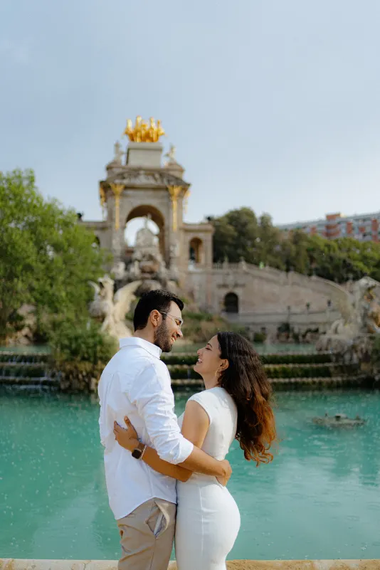 A couple laughs together by the turquoise fountain during a surprise proposal photoshoot at Parc de la Ciutadella.