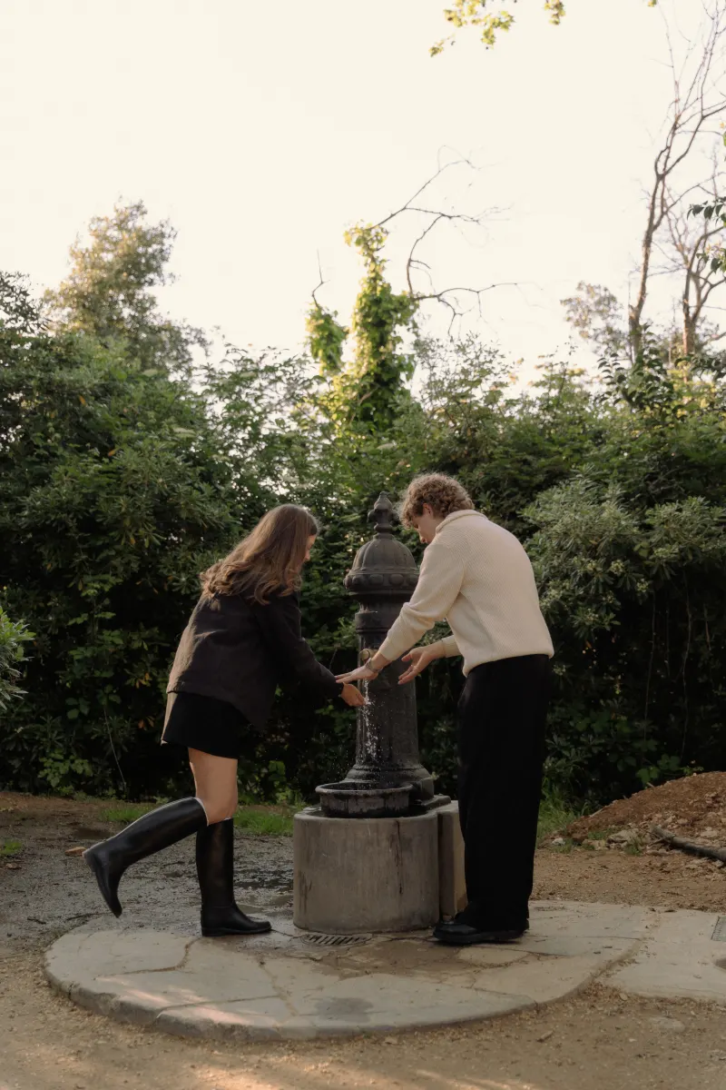 A couple laughs together while washing their hands at a park fountain during a candid engagement photoshoot in Barcelona.