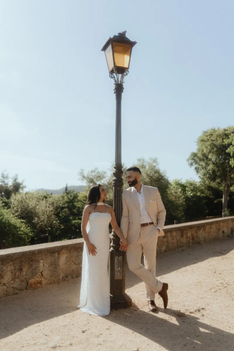 A couple leans against a vintage lamppost, smiling at each other during their surprise proposal photoshoot outdoors.