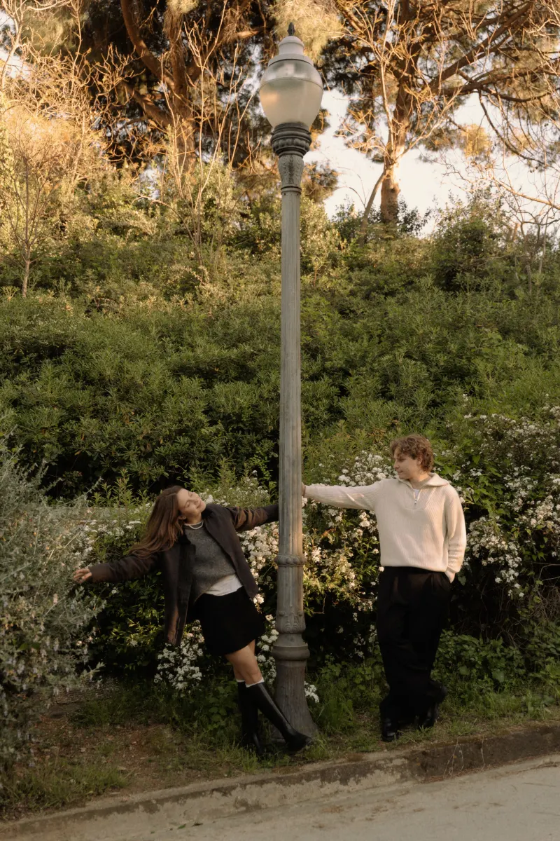 A couple playfully reaches for each other around a lamppost in a sunlit Barcelona park during an engagement photoshoot.