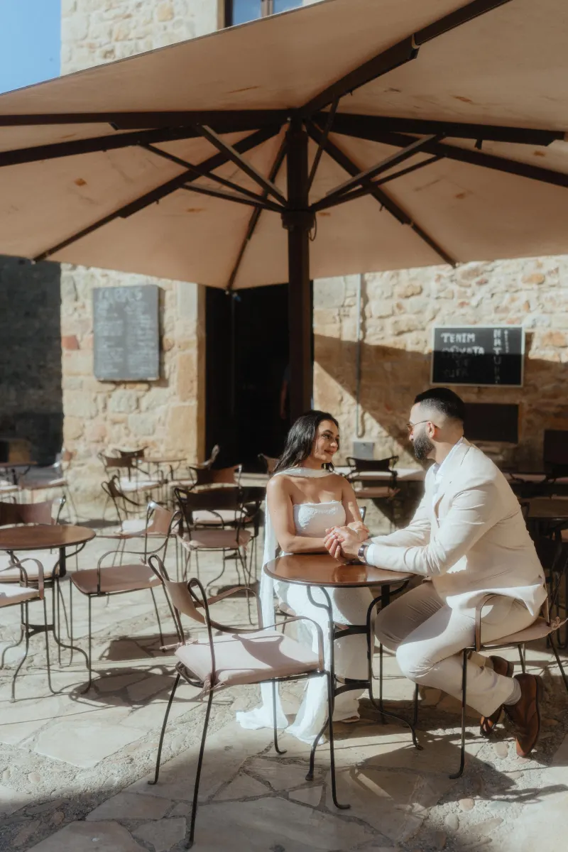 A couple shares a heartfelt moment at a sunlit café table during their surprise proposal photoshoot in a rustic village.