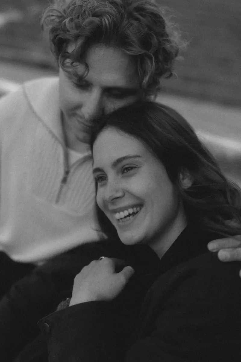 A couple shares a joyful embrace, the woman laughing as her partner gently rests his head beside hers during an engagement sh