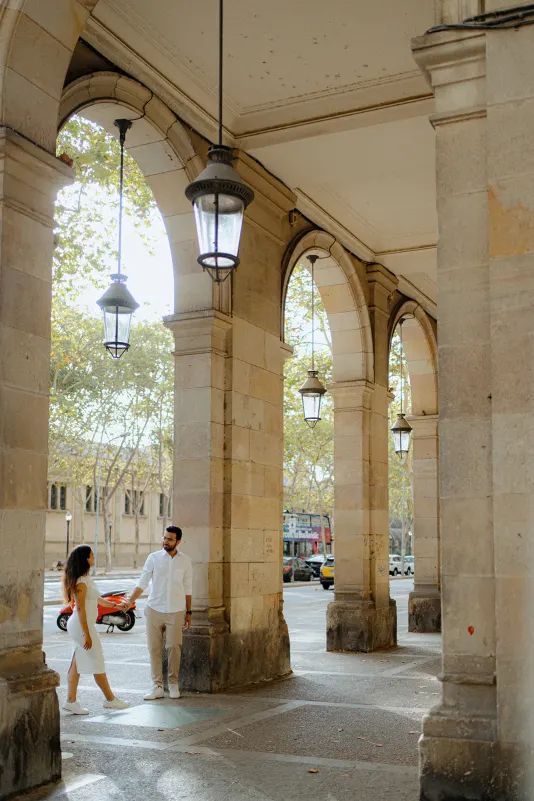 A couple shares a joyful moment under stone arches during a surprise proposal photoshoot in a sunlit city setting.