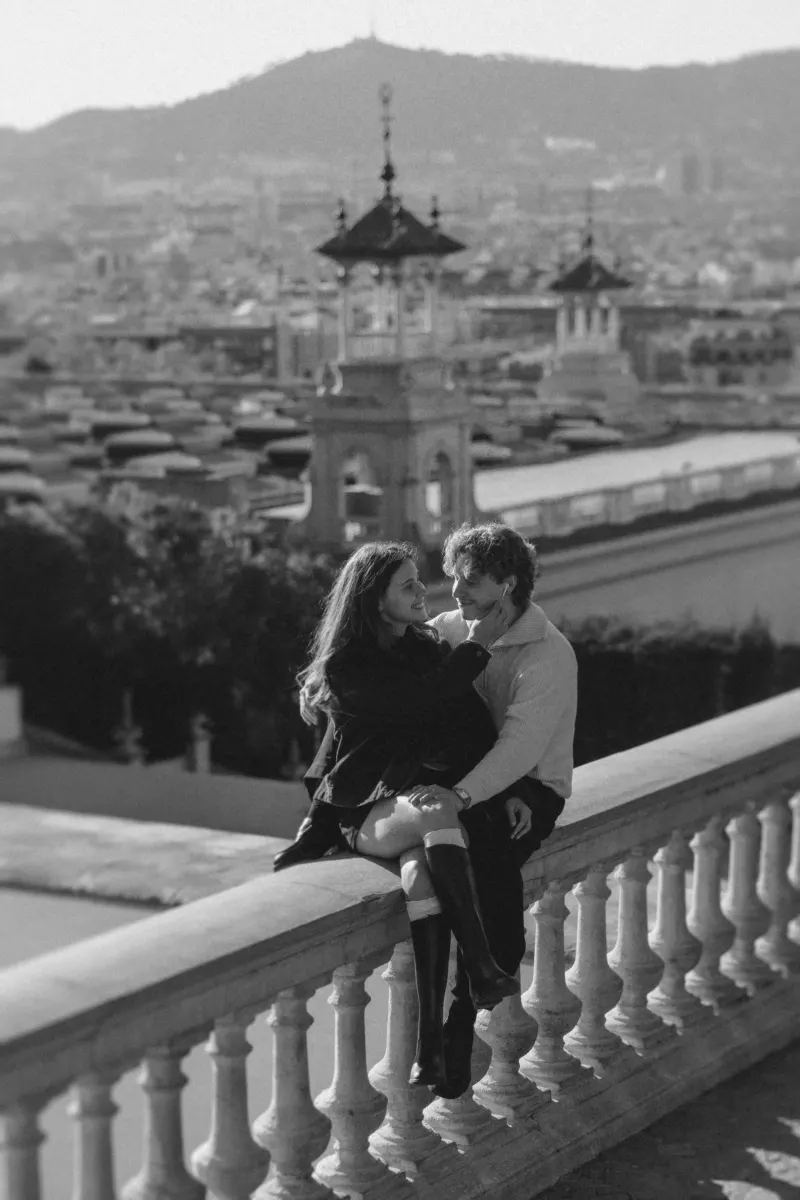 A couple shares a loving gaze while sitting on a stone railing above Barcelona, captured during an engagement photoshoot.