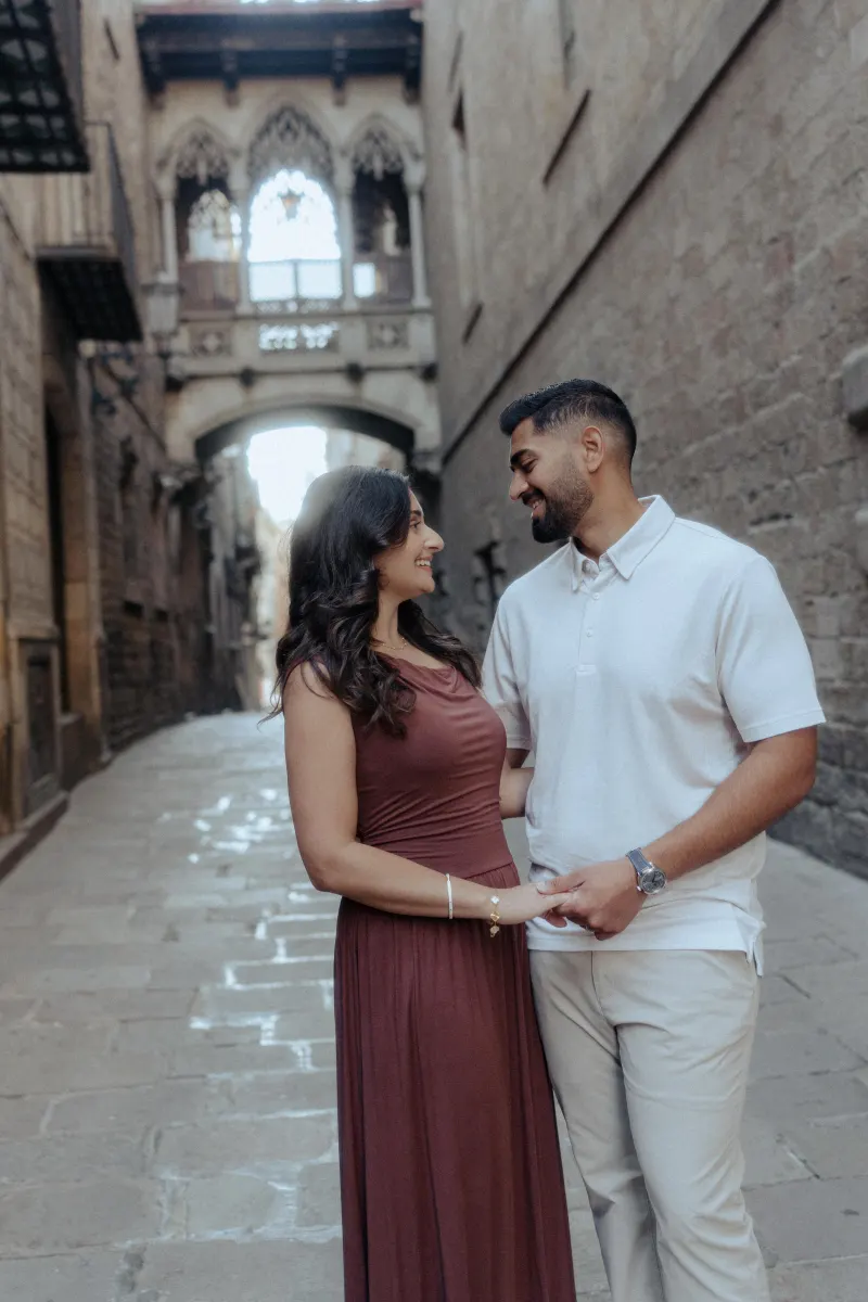 A couple shares a loving smile, holding hands during their engagement photoshoot in a historic European alleyway.