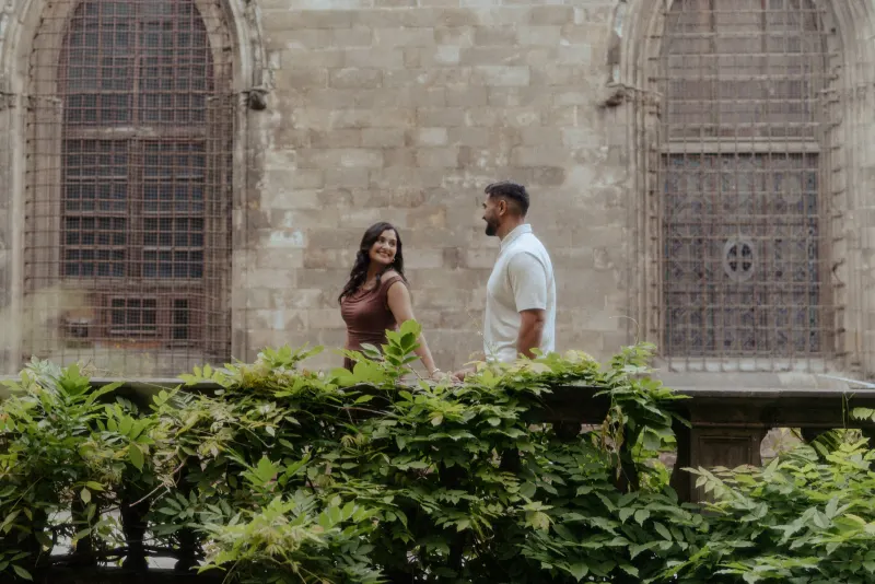 A couple shares a playful glance while walking together during a couples & engagements photoshoot in a leafy courtyard.