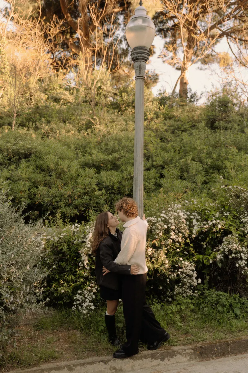 A couple shares a romantic embrace beneath a lamppost in a lush Barcelona park during their engagement photoshoot.