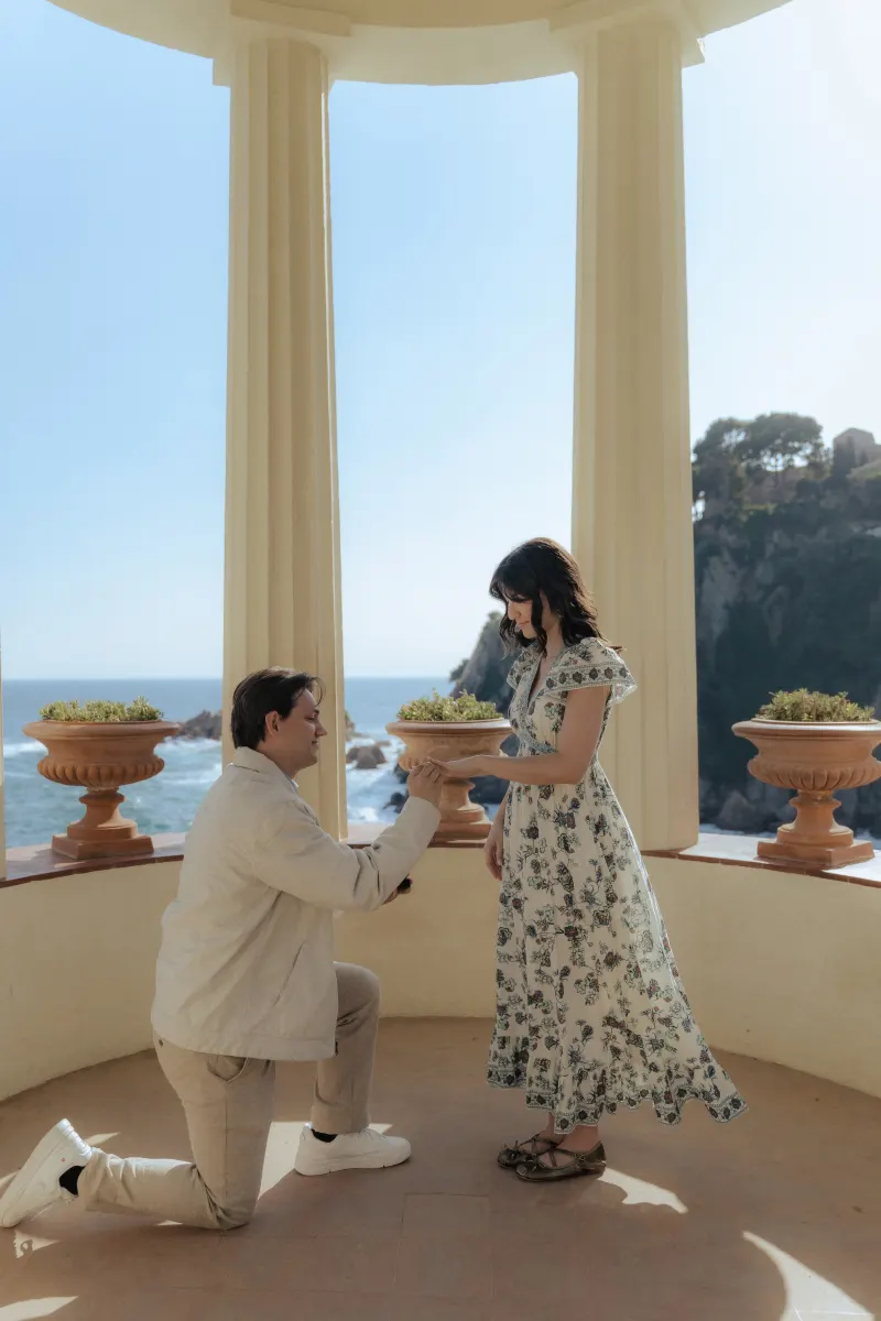 A couple shares a tender moment during a surprise proposal photoshoot by the sea, framed by elegant columns.