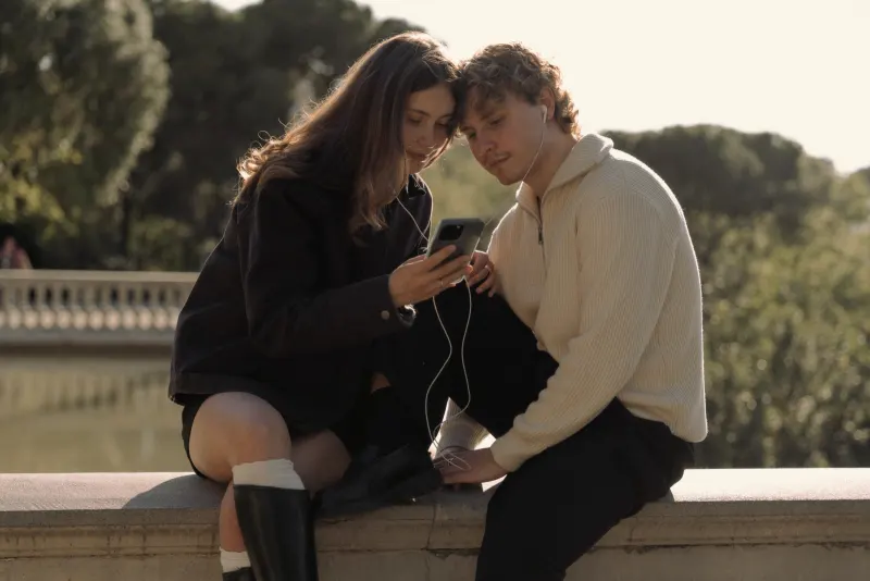 A couple shares headphones and smiles at a phone together on a sunny day in Barcelona during a couples photoshoot.