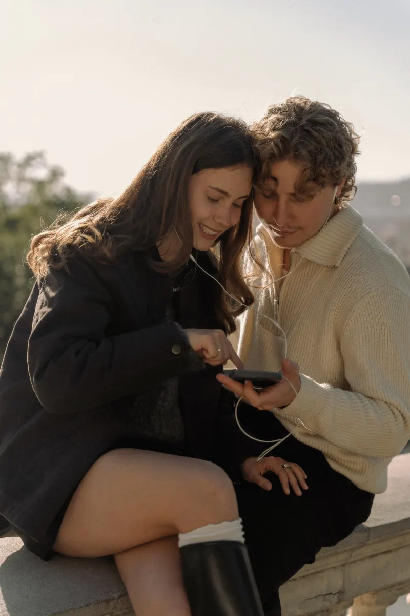A couple shares headphones and smiles at a phone together on a sunny terrace in Barcelona during an engagement session.