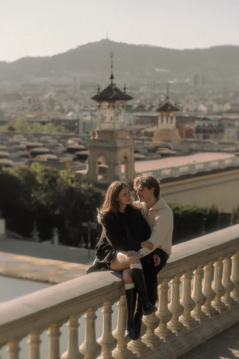 A couple sits arm in arm on a terrace railing in Barcelona, sharing a loving gaze during their engagement photoshoot.