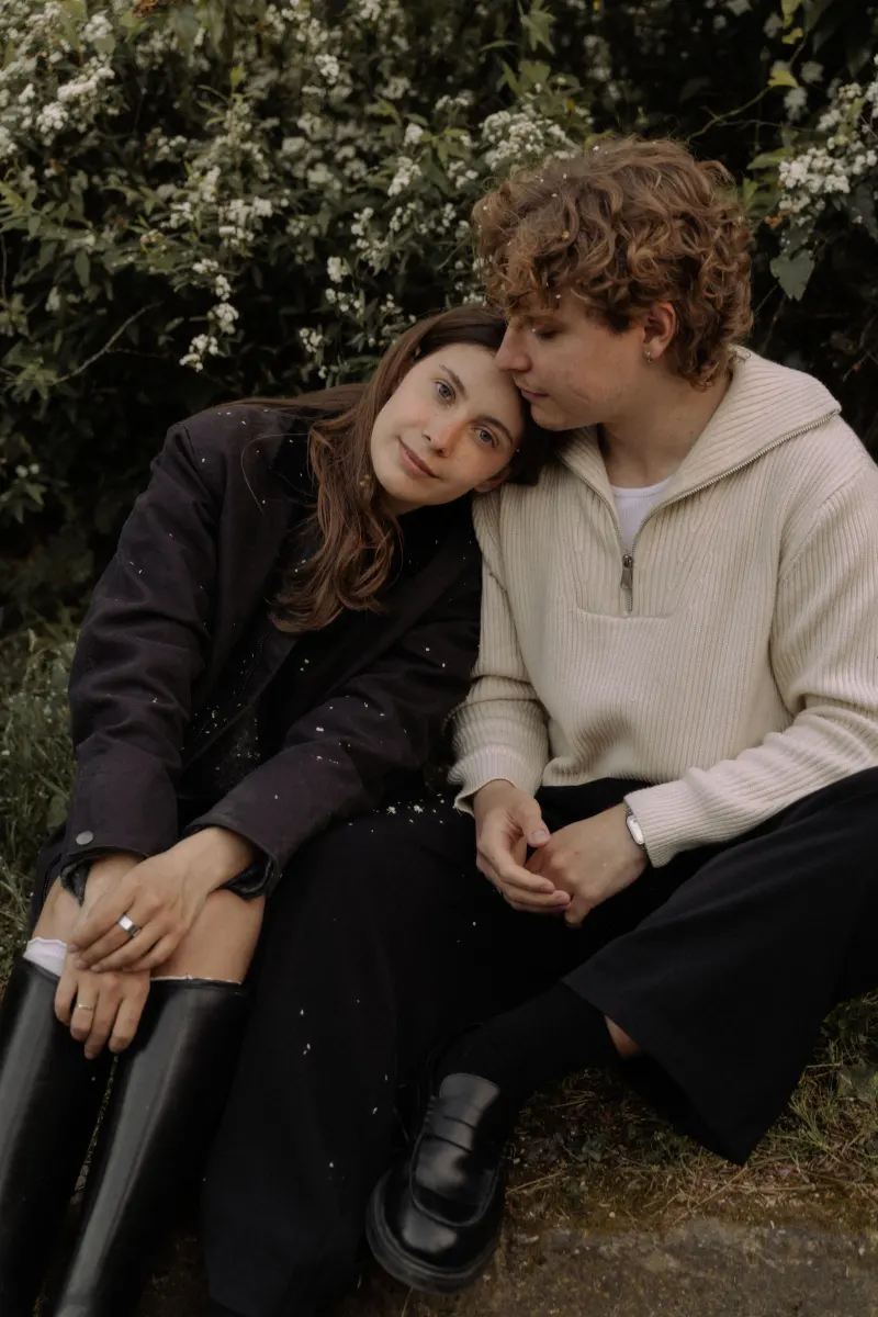 A couple sits close in a garden, the woman resting her head on her partner’s shoulder during an engagement photoshoot.