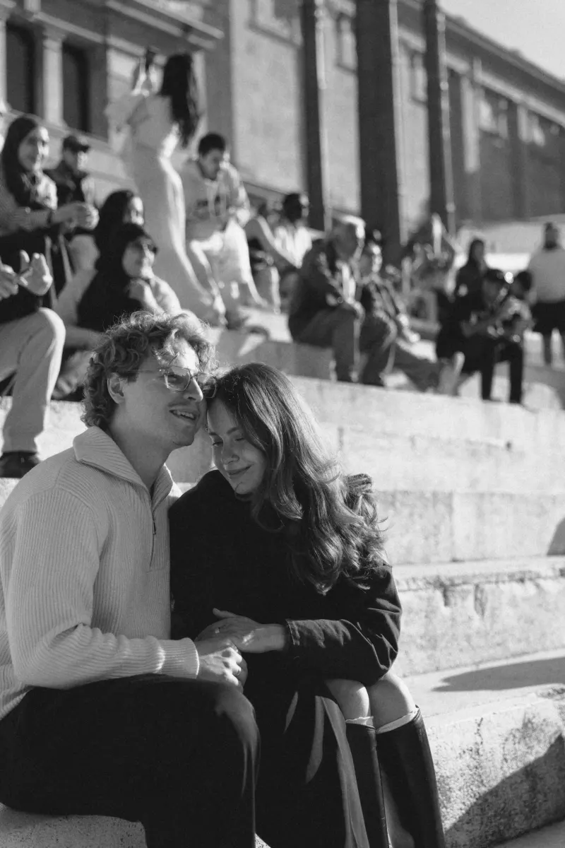 A couple sits close on sunlit steps in Barcelona, sharing a joyful moment during a couples & engagements photoshoot.