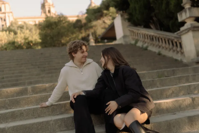 A couple sits close on sunlit steps in Barcelona, sharing a warm smile during their engagement photoshoot.
