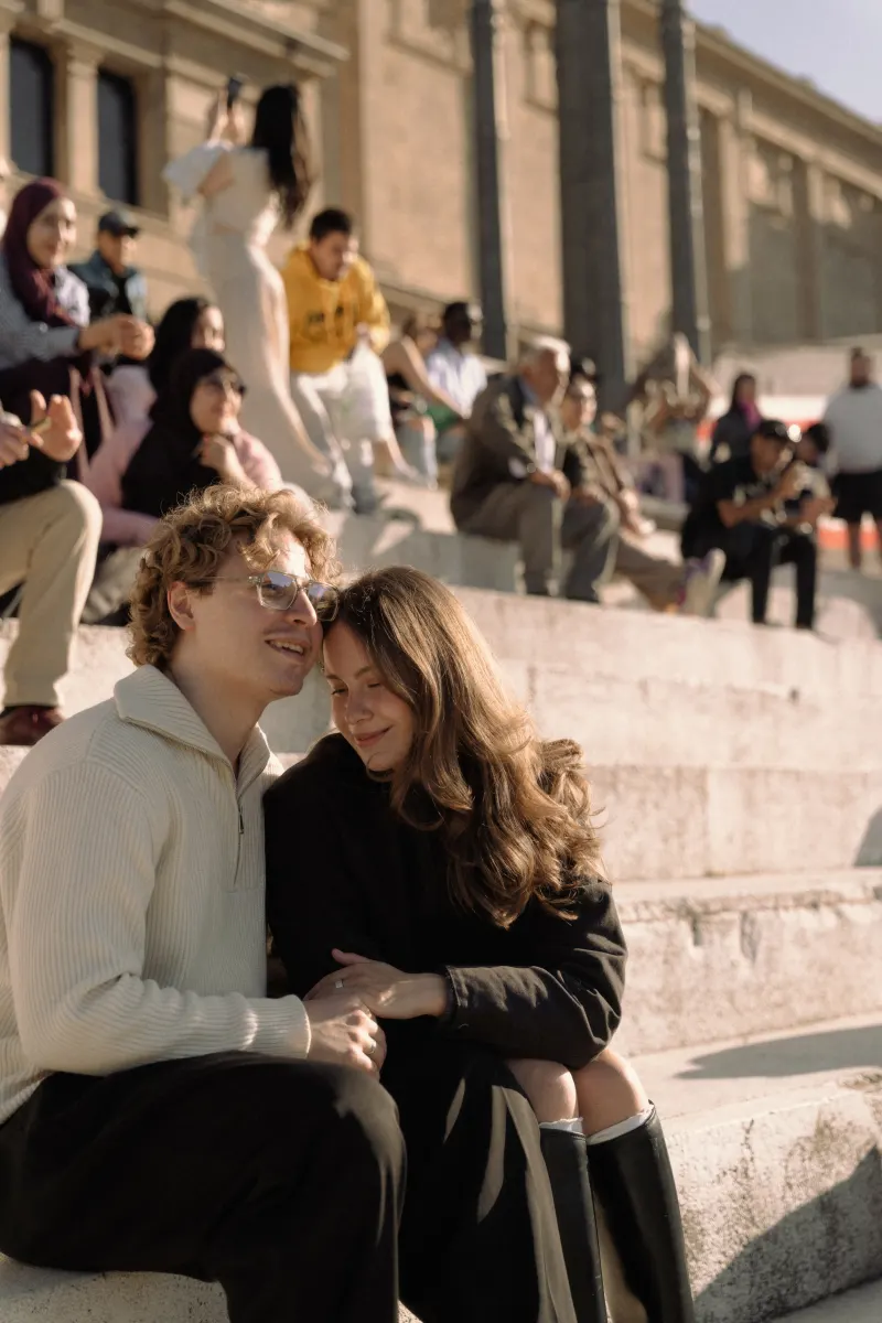 A couple sits close together on sunlit steps in Barcelona, sharing a joyful moment during their engagement session.