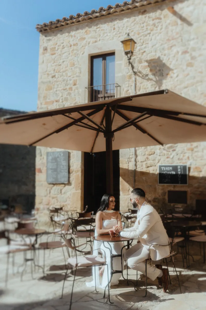 A couple sits hand in hand at a sunlit café table, sharing an intimate moment during their surprise proposal session.