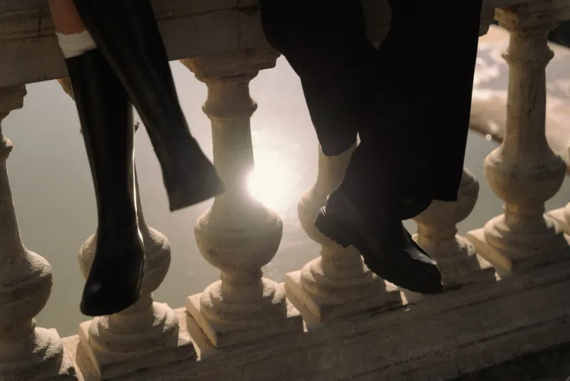 A couple sits side by side on a sunlit stone railing, their legs dangling during a Barcelona engagement photoshoot.