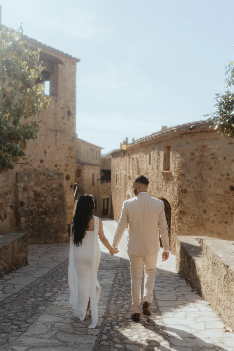 A couple strolls hand in hand through a sunlit stone village, celebrating their surprise proposal photoshoot.