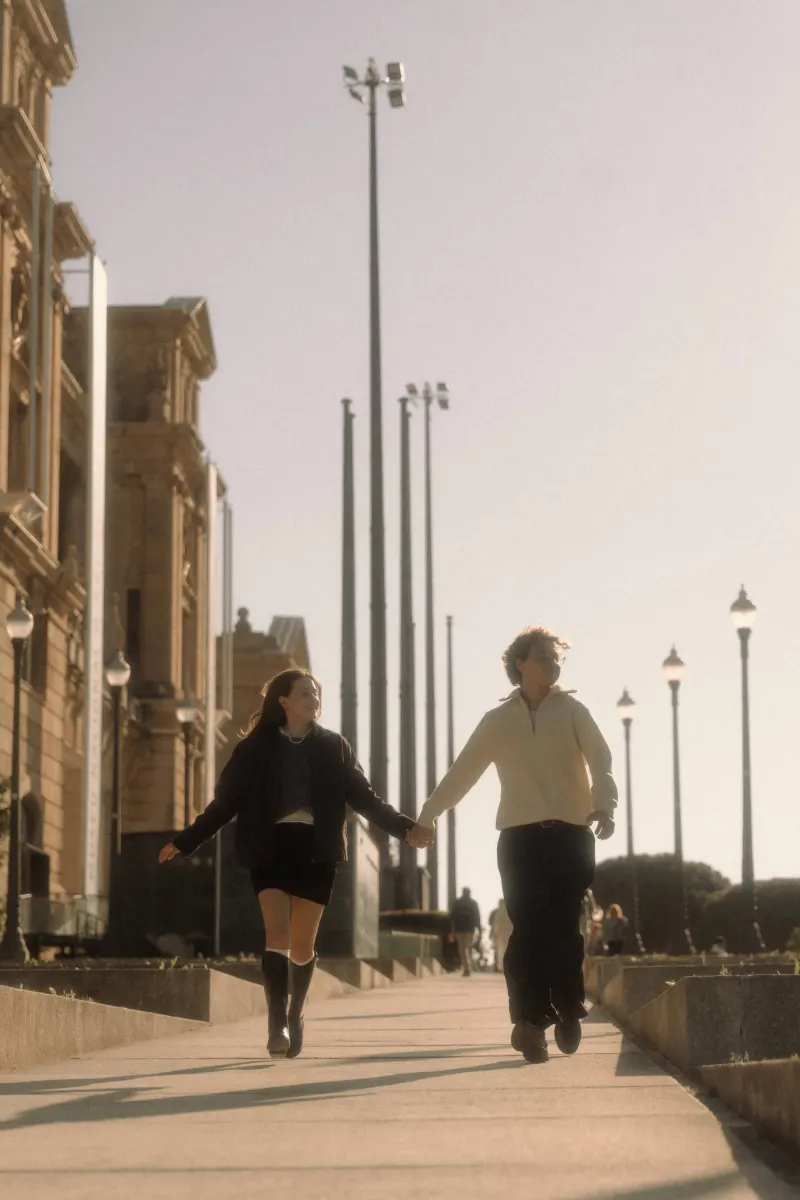 A couple walks hand in hand along a sunlit Barcelona street, sharing a joyful moment during their engagement session.