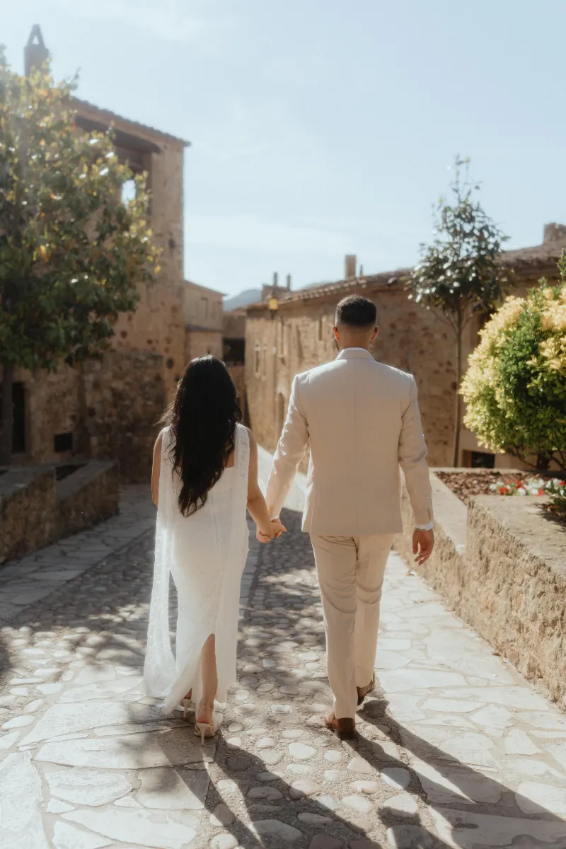 A couple walks hand in hand down a sunlit cobblestone path after their surprise proposal photoshoot in a historic village.