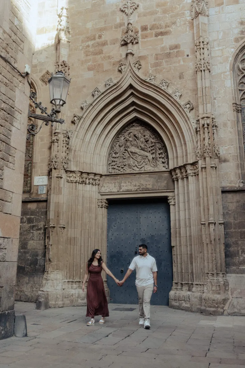 A couple walks hand in hand, smiling at each other during an engagement session by a grand gothic doorway.