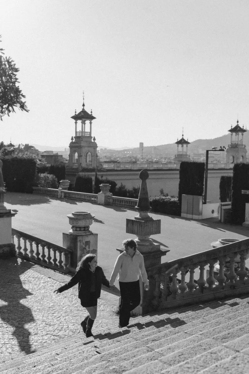 A couple walks hand in hand up sunlit stone steps in Barcelona, smiling during a couples & engagements photoshoot.
