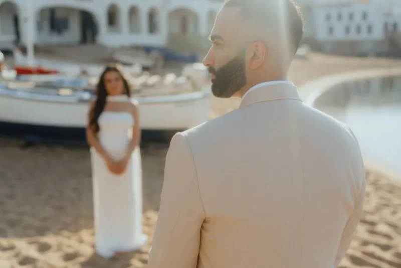 A groom-to-be turns toward his partner on a sunlit beach during a surprise proposal photoshoot.