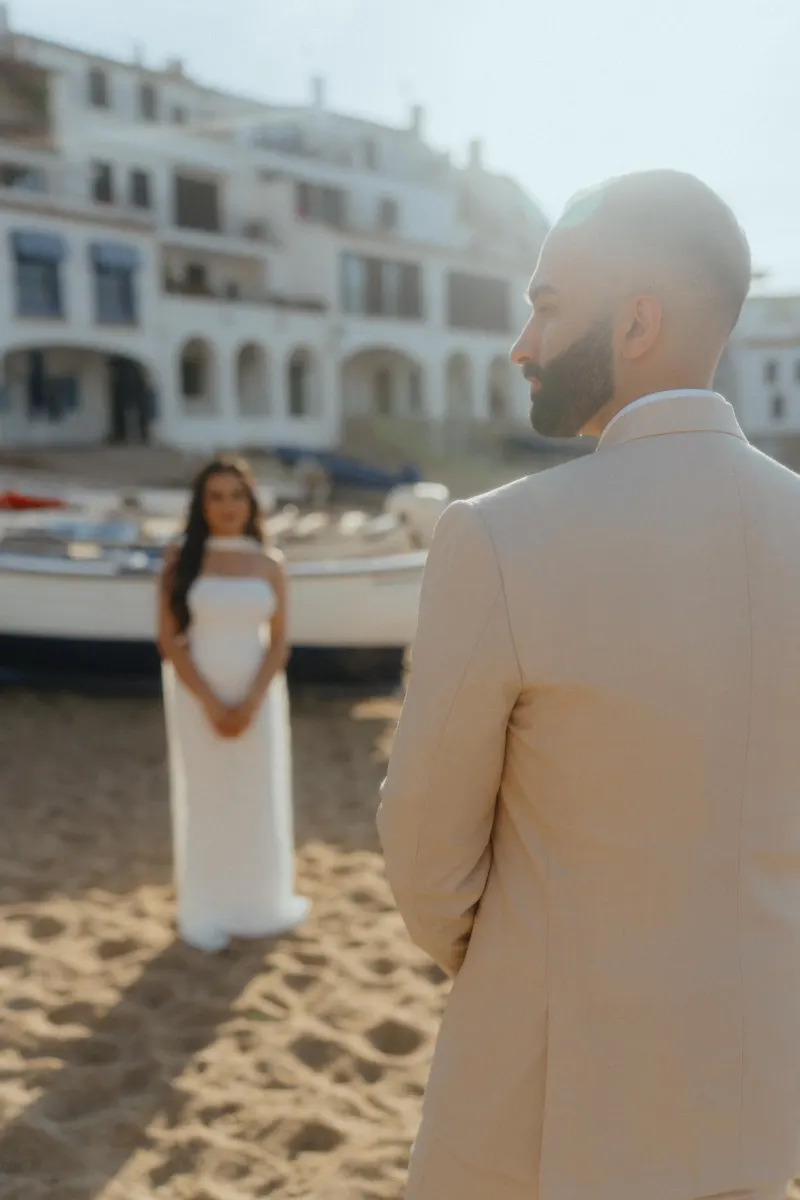 A groom-to-be waits on a sunlit beach as his partner approaches, captured during a surprise proposal photoshoot.