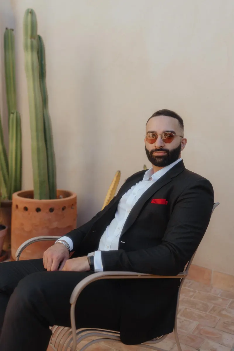A man in a suit with sunglasses sits relaxed on a sunlit patio, awaiting his surprise proposal moment.