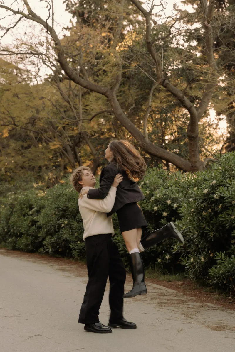 A man lifts his partner in a joyful embrace on a tree-lined path during a couples & engagements photoshoot in Barcelona.