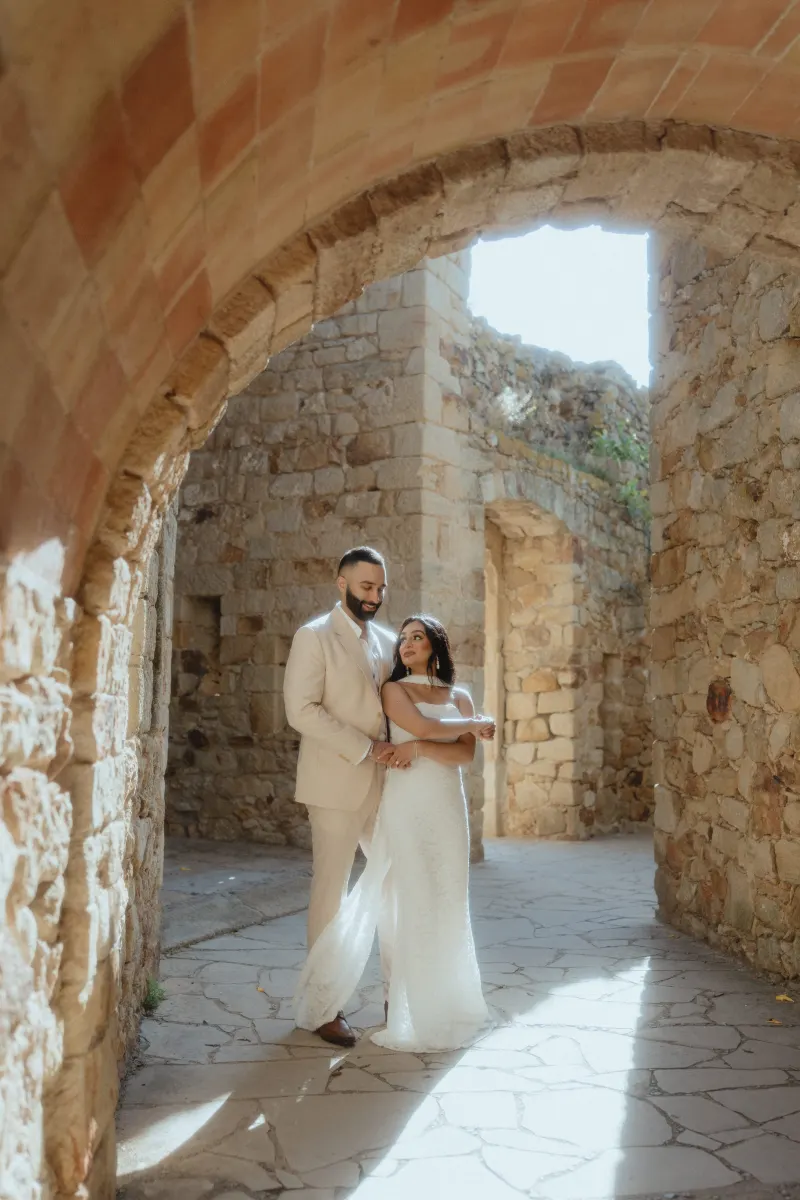 A newly engaged couple embraces in soft sunlight beneath a rustic stone archway during their surprise proposal photoshoot.