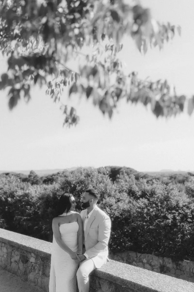 A newly engaged couple sits close on a stone wall, sharing a tender look during their surprise proposal photoshoot.